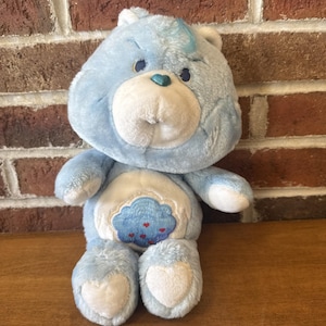 Może przedstawiać: Jasnoniebieski pluszowy miś z białym brzuchem i stopami w kształcie serca. Miś ma wzór chmury na brzuchu z czerwonymi sercami. Ma jasnoniebieski pas włosów i małe niebieskie serce na nosie.