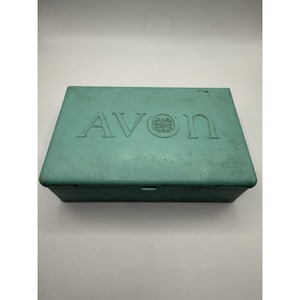 Puede incluir: Una caja rectangular de color turquesa con la palabra "AVON" en relieve en la tapa. La caja tiene un diseño sencillo y parece estar hecha de un material sólido. El logo de Avon es visible entre las letras V y N.