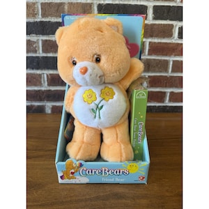Puede incluir: Un peluche naranja de Care Bear, Friend Bear, en su caja original. El oso tiene una panza blanca con un diseño floral. La caja presenta el logotipo de Care Bears y las palabras "Friend Bear". El juguete es nuevo y en su embalaje original.