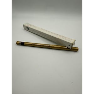 May include: A gold-colored eyeliner pencil with a dark purple band and text. The pencil is next to its white and gold box, which has the brand name "Mary Kay" and other text.