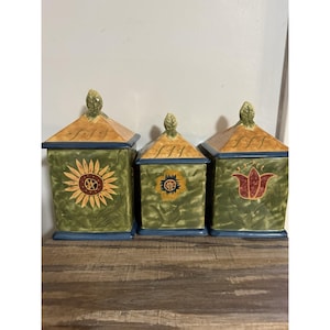 May include: A set of three ceramic kitchen canisters with lids. Each canister is square with a green base and sides, blue trim, and a tan roof. They are decorated with a sunflower, a flower, and a tulip.