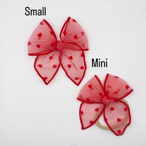 May include: Two red hair bows with heart patterns. The larger bow is labelled "Small" and the smaller bow is labelled "Mini". Both bows have a sheer, airy texture with red trim and small red hearts scattered across the fabric. The mini bow is attached to a beige headband.