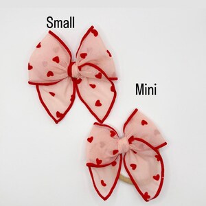 May include: Two pink hair bows with red heart patterns and red trim are shown against a white backdrop. The larger bow is labelled "Small," and the smaller bow is labelled "Mini." The bows are made of a sheer fabric.