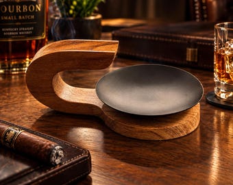 May include: A cigar rest and ash tray combination featuring a curved, natural-toned wooden base with a smooth grain texture. A circular, matte black metal tray is inset into the wooden structure, providing a stable surface for ash collection.
