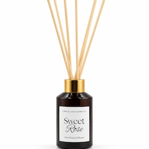 Peut inclure: Un flacon diffuseur à roseaux en verre ambré foncé avec un col doré et six bâtonnets en bois clair. L'étiquette blanche porte le texte Lilies & Lace Candle Co., Sweet Rose et 110ml Reed Diffuser en polices serif et script noires.