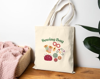 Pode incluir: Saco tote creme com as palavras "Sewing Bag" em verde. O saco apresenta um gráfico de materiais de costura, incluindo carretéis de linha, tesouras, alfinetes, botões e alfinetes de segurança. O saco tem uma alça e é feito de um material semelhante a lona.