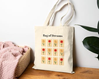 May include: Cream-colored tote bag with the text "Bag of Dreams" and a pattern of stylized flowers in red and yellow. The bag has long handles and is made of a sturdy fabric, suitable for carrying groceries or other items.