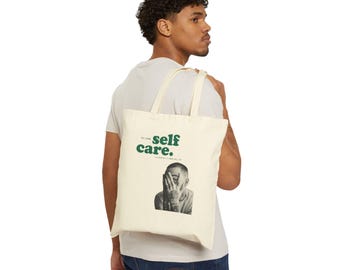 May include: Cream tote bag featuring the words "self care" in green, and a black and white image of a person covering their face. The bag has cream straps and is carried over the shoulder.