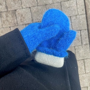 Könnte beinhalten: Ein Paar leuchtend blaue Fäustlinge mit hellgrauem Strickbündchen. Die Fäustlinge sind aus einem weichen, flauschigen Material gefertigt. Die Fäustlinge werden vor einem dunkelblauen Mantel und einem grauen Ziegelhintergrund gezeigt.