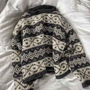 Könnte beinhalten: Ein langärmeliger Pullover mit Rundhalsausschnitt und einem geometrischen Muster in Dunkelanthrazit und Creme. Das Strickkleidungsstück zeigt wiederkehrende horizontale Bänder mit komplexen Rauten- und Kreuzmotiven auf Brust, Ärmeln und Saum.
