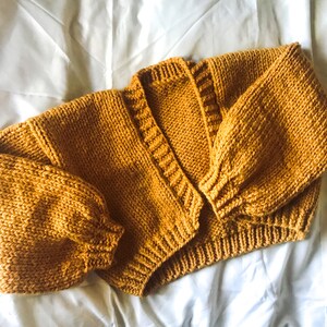 Könnte beinhalten: Ein senfgelber Strickcardigan mit langen Ärmeln und V-Ausschnitt.
