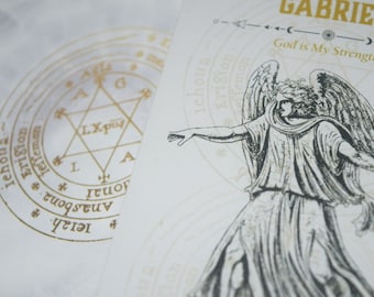 May include: Two white cards with gold-colored illustrations. One card features a detailed drawing of an angel with outstretched arms. The other card has a circular design with a star and text. The word "GABRIEL" is at the top of the angel card.