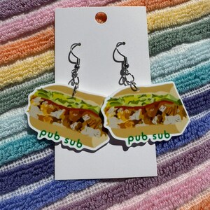 Puede incluir: Un par de pendientes novedosos con forma de sándwich. Los pendientes tienen un fondo blanco con una colorida ilustración de un sándwich y las palabras "pub sub" en verde. Cuelgan de ganchos plateados.