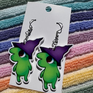 May include: A pair of dangle earrings featuring green cartoon creatures wearing purple witch hats. The earrings have silver-colored fish hook closures and are displayed on a white card against a colorful striped background.