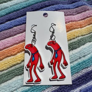 May include: A pair of novelty earrings featuring a red figure with a blue tie. The earrings are attached to silver-colored hooks and are displayed on a white card against a striped background.