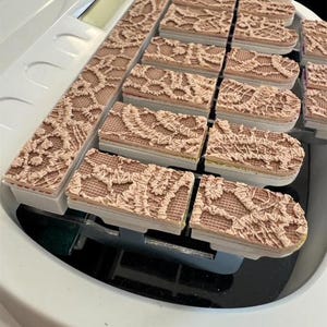 May include: A collection of rectangular stamps with a detailed lace pattern in a light brown color. The stamps are arranged in rows and columns, resting on a white surface with a digital display.