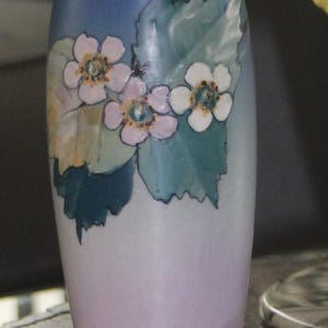 May include: A ceramic vase with a blue top and a light purple base. The vase is decorated with painted flowers in shades of pink and white, with green leaves. The vase is approximately 8 inches tall.