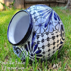 May include: A white ceramic pot with blue and white floral designs. The pot is decorated with a grid pattern and has a wide opening at the top. The pot is lying on its side on a bed of green grass.