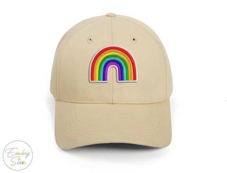Op de afbeelding: Beige baseballpet met een kleurrijke regenboogpatch. De regenboog heeft de kleuren rood, oranje, geel, groen, blauw en paars. De pet heeft een gebogen klep en een knoop aan de bovenkant.