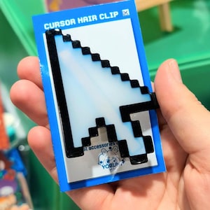 May include: A pixelated computer mouse cursor hair clip with a white centre and a thick black outline. The hair clip is attached to a rectangular blue and white display card that features the text CURSOR HAIR CLIP and a small logo with the word YORUM.