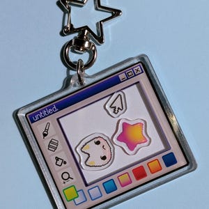 Op de afbeelding: Een doorzichtige acryl sleutelhanger in de vorm van een computervenster met de titel untitled. Het ontwerp toont een digitaal tekeninterface met een penseel, gum, vergrootglas, een pixelachtig kattengezicht, een ster en een cursor.