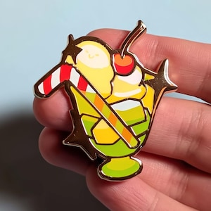 May include: A gold-colored enamel pin in the shape of a sundae. The sundae features yellow ice cream, a red cherry, a candy cane, and a green base. The pin has a shiny finish and is set against a light blue background.