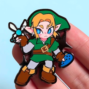 May include: Enamel pin depicting a cartoon character in a green tunic and hat, brown boots, and blonde hair. The character holds a blue object and a white and blue fairy. The pin has a black outline and a gold buckle.