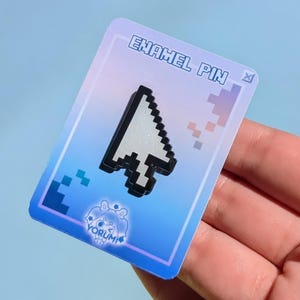 May include: A white and black pixelated computer mouse cursor enamel pin attached to a rectangular backing card. The card features a blue and purple gradient, the text ENAMEL PIN at the top, and a small YORUMI logo with a character graphic.