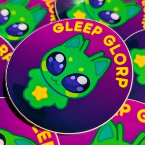 May include: A collection of round stickers featuring a cartoon alien character. The alien is green with large blue eyes, a yellow star on its chest, and the words "GLEEP GLORP" in yellow text around the edge of the purple background.