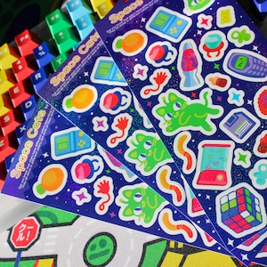 May include: Three sheets of holographic stickers with a space cat theme. The stickers feature a variety of designs, including cats, planets, stars, and other space-themed imagery. The stickers are printed on a blue background with a glittery finish.
