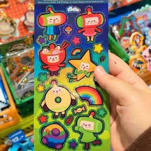 May include: A sheet of colorful stickers featuring cartoon characters with the text "ROLLING STARS COUSINS" and "www.yorumi.art". The stickers include various shapes and designs, such as stars, rainbows, and food items. The background is a vibrant green.