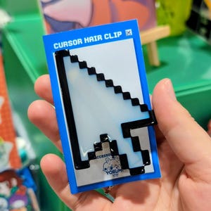 May include: A white and black pixelated computer mouse cursor hair clip attached to a blue rectangular display card. The card features the text CURSOR HAIR CLIP at the top and a small logo with the word YORUMI at the bottom centre.