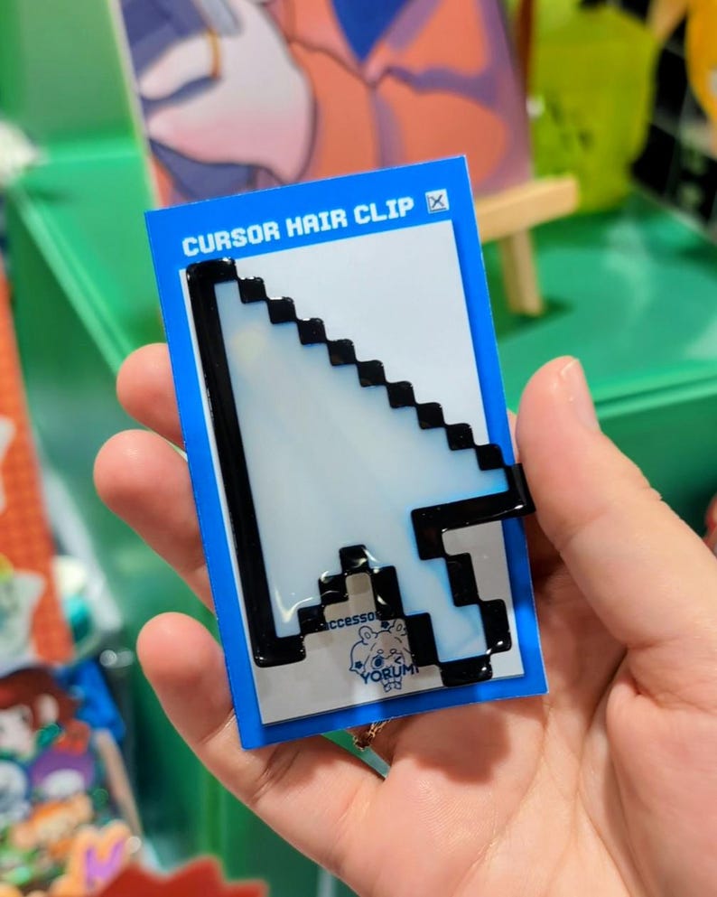 May include: A white and black pixelated computer mouse cursor hair clip attached to a blue rectangular display card. The card features the text CURSOR HAIR CLIP at the top and a small logo with the word YORUMI at the bottom centre.