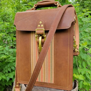 May include: A brown leather briefcase with a striped fabric lining and a brass buckle. The bag has a long adjustable strap and a top handle.