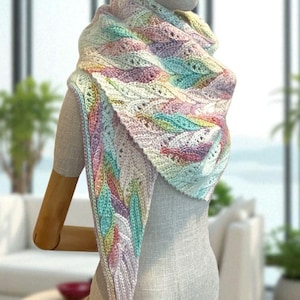 May include: A pastel-colored, knitted shawl displayed on a mannequin. The shawl features a repeating leaf pattern in shades of pink, blue, green, and yellow. The design is intricate, with openwork details. The shawl is draped over the mannequin's shoulders.