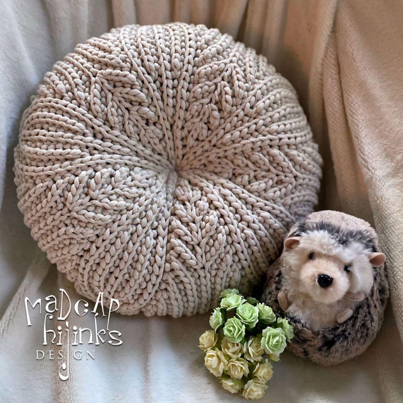 May include: A beige knitted round cushion with a textured pattern, a small stuffed hedgehog, and a bouquet of green and white flowers. The cushion has the text "Madcap Hijinks Design" on the bottom.