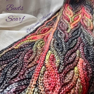 Puede incluir: Una bufanda de ganchillo colorida con un patr&oacute;n texturizado. La bufanda est&aacute; hecha con una variedad de tonos de hilo rojo, naranja, amarillo, verde y gris. El texto "Autumn Buds Scarf by Yvonne Martinez" est&aacute; escrito en la bufanda.