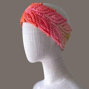 Puede incluir: Una diadema de ganchillo con un degradado de hilo naranja, rosa y amarillo en un patrón de hojas. El texto "The Beauty Crown" y "MajesticArtCrochet" es visible en la parte superior. La diadema se muestra sobre un maniquí blanco texturizado.