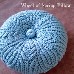 May include: A light blue crocheted pillow with a button in the centre. The pillow is round and has a textured surface. The text "Wheel of Spring Pillow" is written above the pillow.