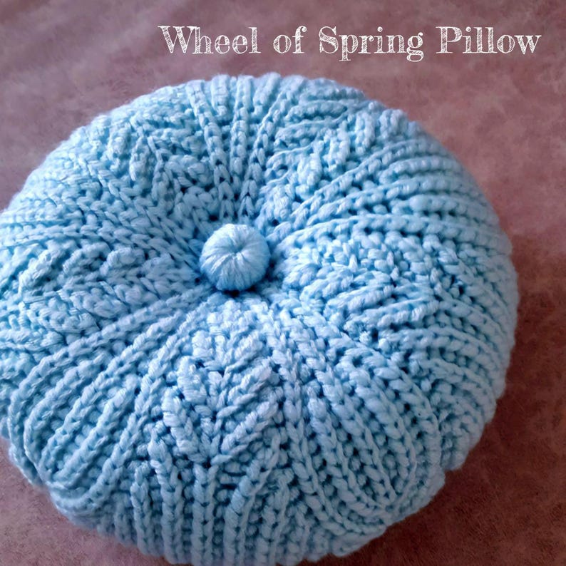 May include: A light blue crocheted pillow with a button in the centre. The pillow is round and has a textured surface. The text "Wheel of Spring Pillow" is written above the pillow.