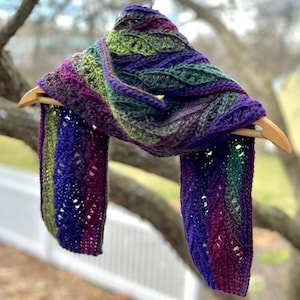 May include: A crocheted scarf with a leafy pattern in shades of purple, green, and brown. The scarf is hanging on a wooden hanger.