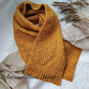 Puede incluir: Una bufanda de crochet de color amarillo mostaza con un patr&oacute;n de punto de cable. La bufanda est&aacute; doblada por la mitad y colocada sobre una superficie blanca.