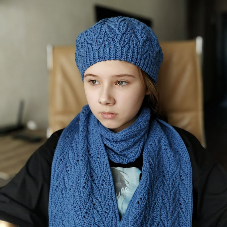 May include: A blue knitted beret and matching scarf. The beret has a cable knit pattern and the scarf is a long, rectangular shape.