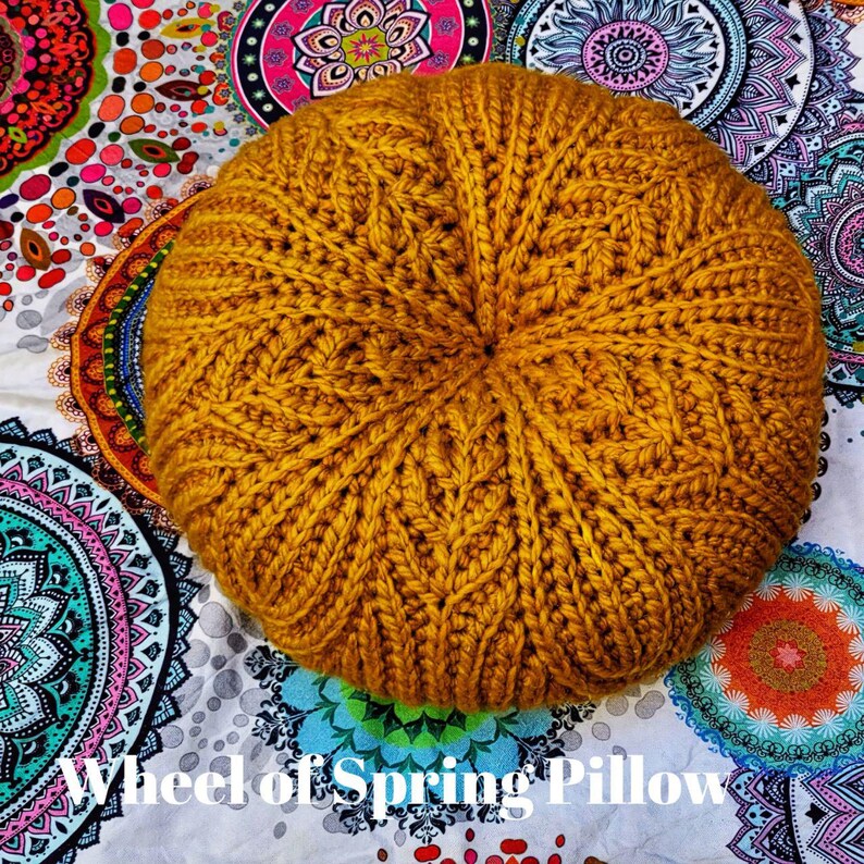 May include: A round, mustard yellow crocheted cushion with a cable knit pattern. The cushion is sitting on a white fabric with colourful mandala designs.