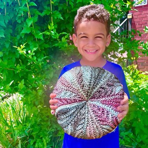 May include: A young person holding a large, round, crocheted cushion with a multi-coloured pattern of pink, brown, and white.