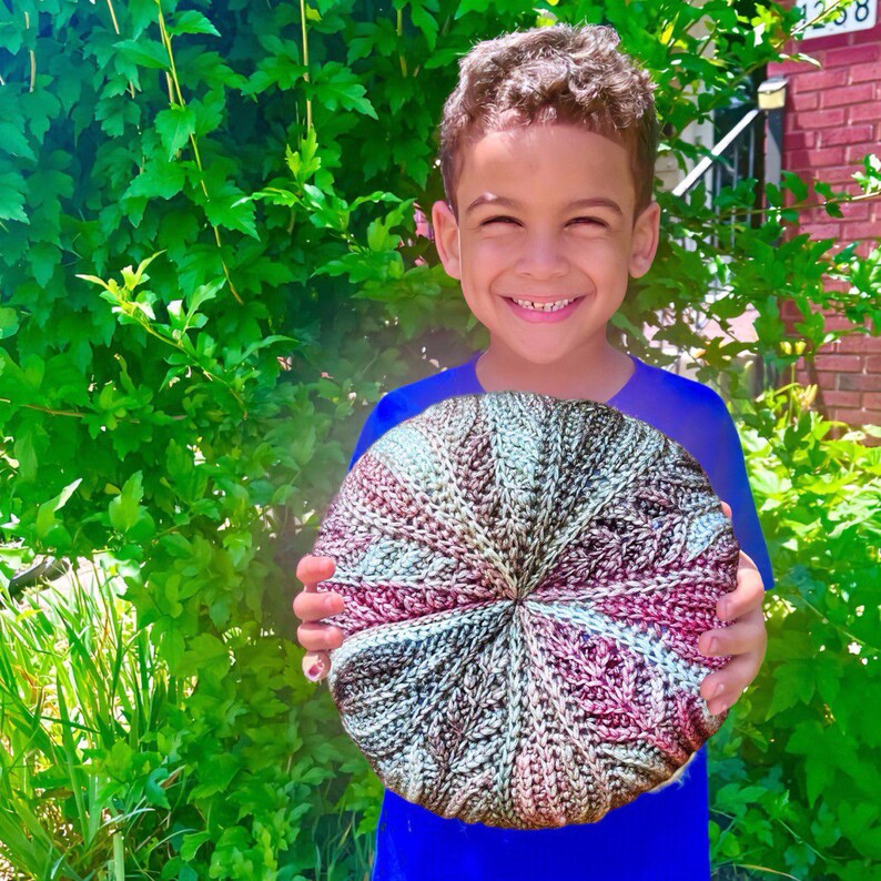 May include: A young person holding a large, round, crocheted cushion with a multi-coloured pattern of pink, brown, and white.