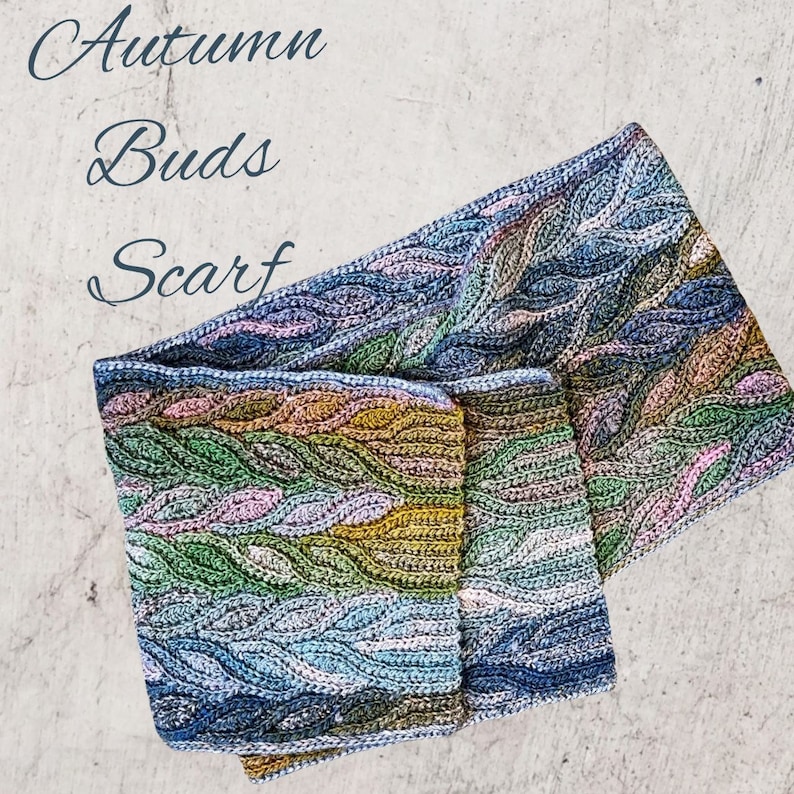 Puede incluir: Una bufanda de ganchillo con un patr&oacute;n de hojas en tonos de azul, verde, amarillo y rosa. La bufanda est&aacute; doblada por la mitad y el texto "Autumn Buds Scarf" est&aacute; escrito encima.