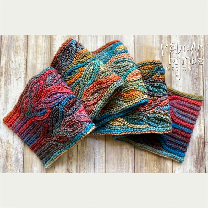 Puede incluir: Una bufanda de ganchillo colorida con un patr&oacute;n de hojas. La bufanda est&aacute; doblada en tres partes y est&aacute; hecha con una variedad de tonos de azul, verde, naranja y rojo. La bufanda est&aacute; sobre una superficie de madera. "Madcap Hijinks Design" est&aacute; escrito en texto blanco sobre fondo negro en la esquina superior derecha de la imagen.