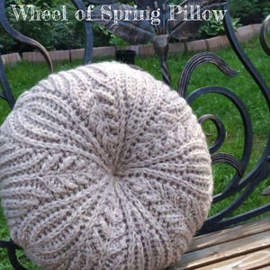 May include: A round, beige crocheted cushion with a cable knit pattern. The cushion is sitting on a wooden bench.