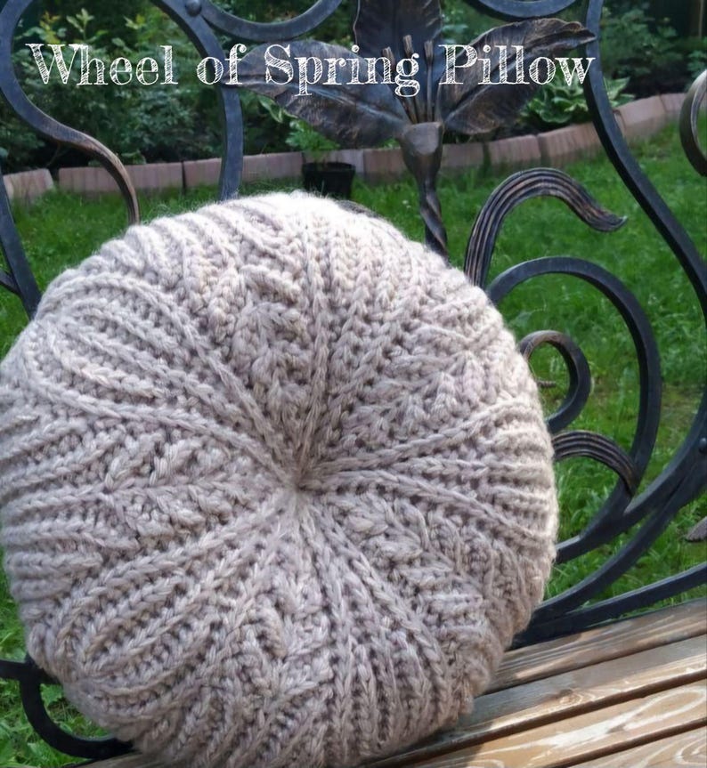 May include: A round, beige crocheted cushion with a cable knit pattern. The cushion is sitting on a wooden bench.